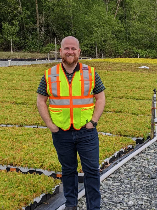 An image of a man standing on a roof with vegetation in the background. He is smiling and wearing a bright hi-visibility vest.