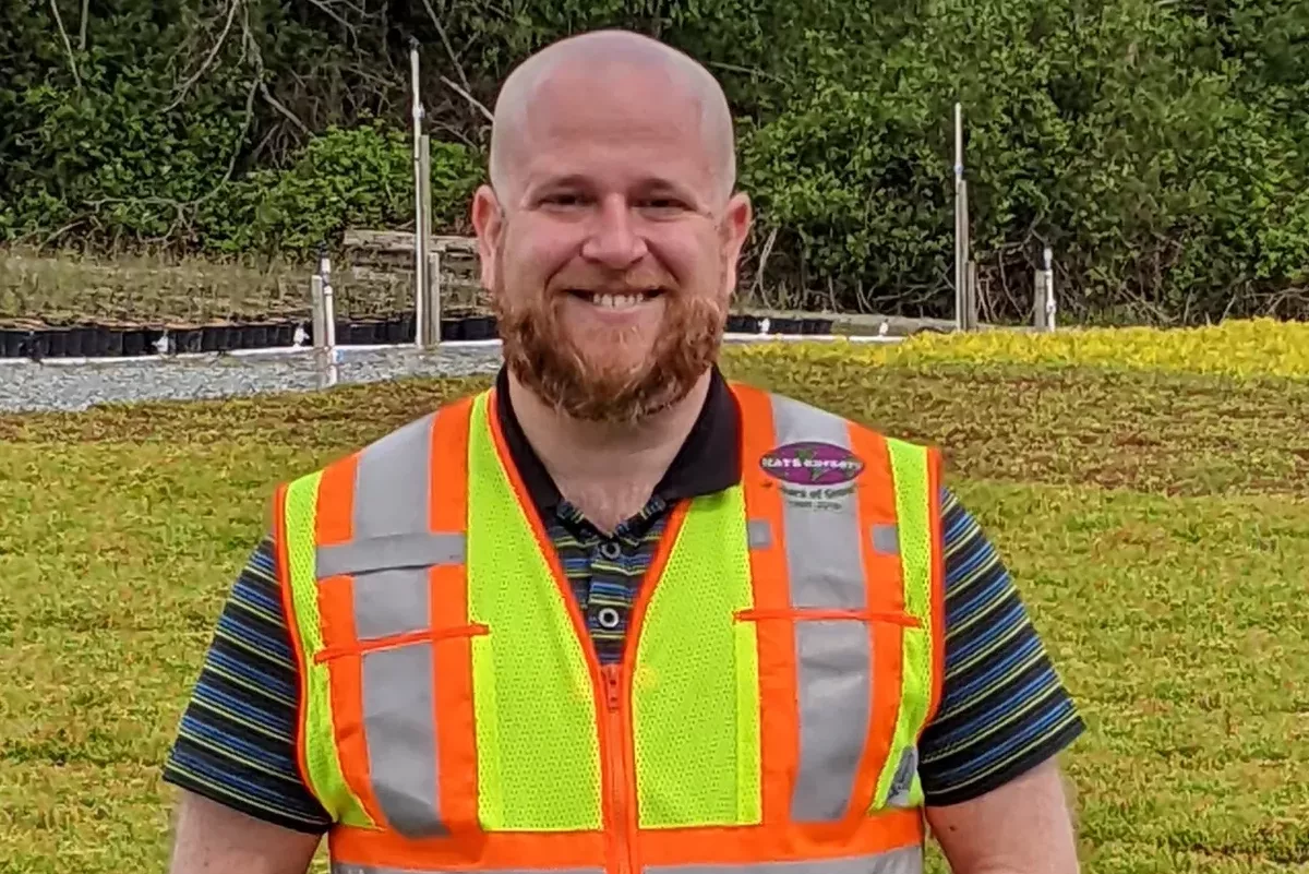 An image of a man standing on a roof with vegetation in the background. He is smiling and wearing a bright hi-visibility vest.